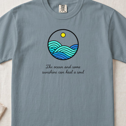 A grey t-shirt featuring a circular graphic of blue ocean waves and a yellow sun with the text "The ocean and some sunshine can heal a soul".