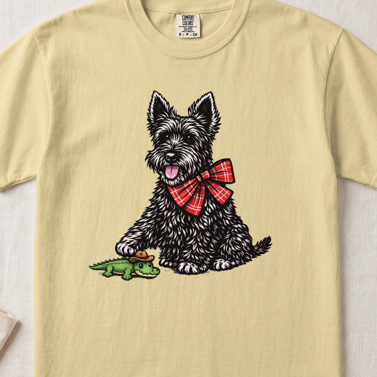 A light yellow t-shirt features a graphic of a black terrier wearing a red plaid bow, touching a toy alligator.