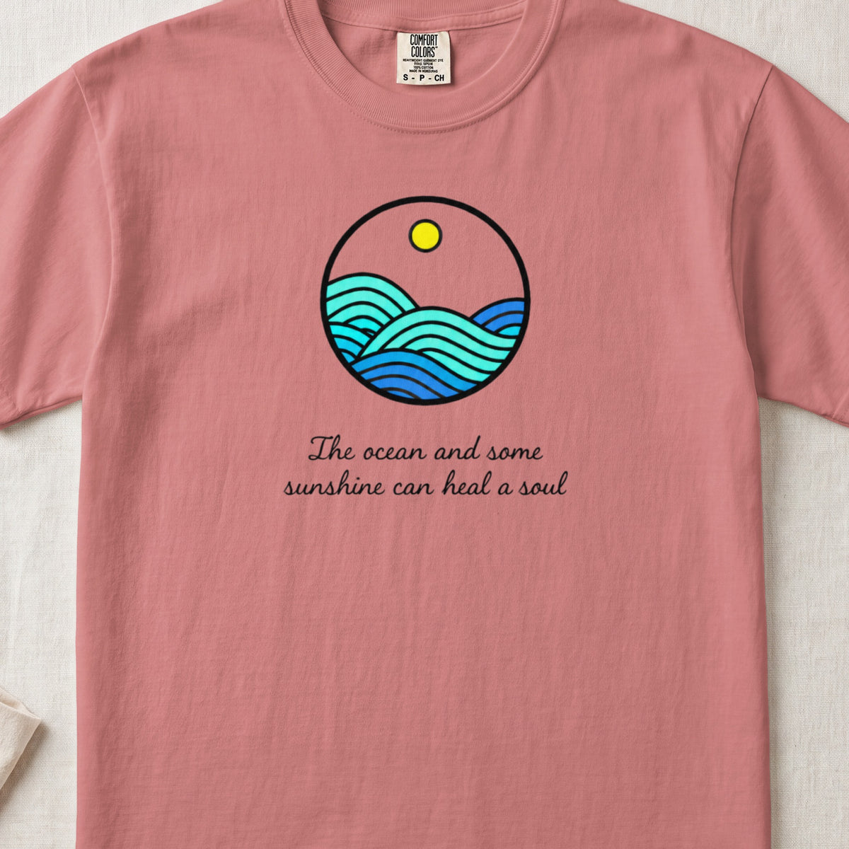 Pink t-shirt featuring a circular ocean graphic with waves, a sun, and the text The ocean and some sunshine can heal a soul.
