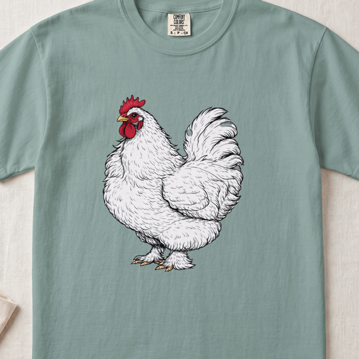 A sage green t-shirt featuring a detailed black-and-white sketch of a fluffy white rooster with a red comb.