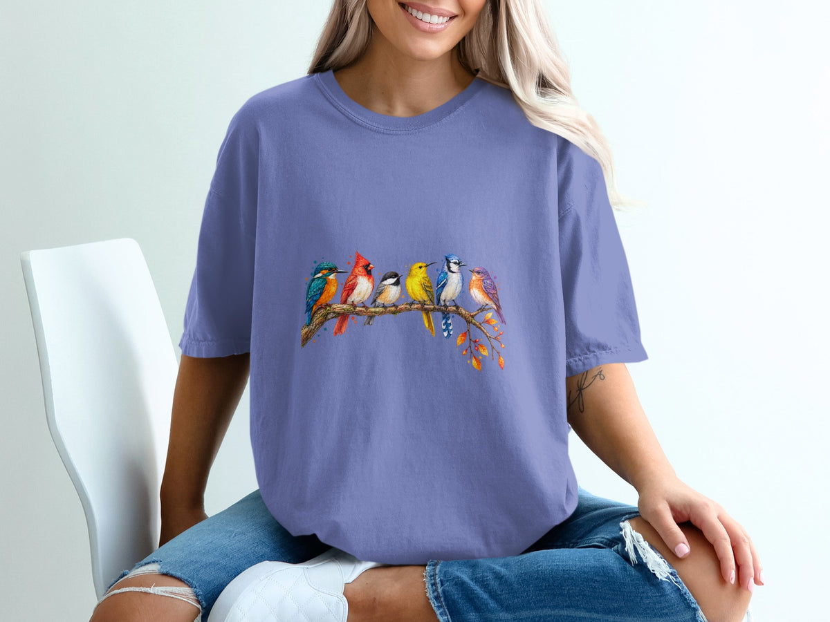 A watercolor painting of colorful birds perched on a tree branch adorns a periwinkle t-shirt.