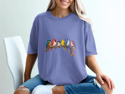 A watercolor painting of colorful birds perched on a tree branch adorns a periwinkle t-shirt.