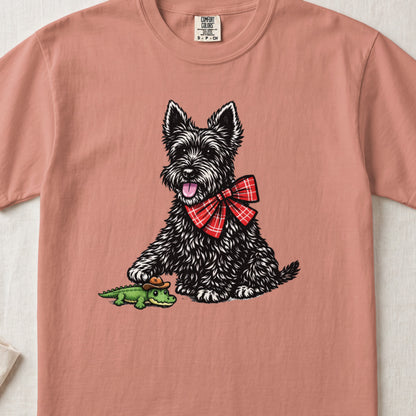 Salmon pink t-shirt featuring a graphic of a black terrier wearing a red plaid bow tie and touching a toy alligator.