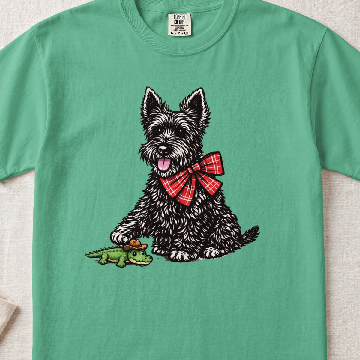 A green t-shirt printed with a black Scottish Terrier wearing a red plaid bow tie and touching a toy alligator.