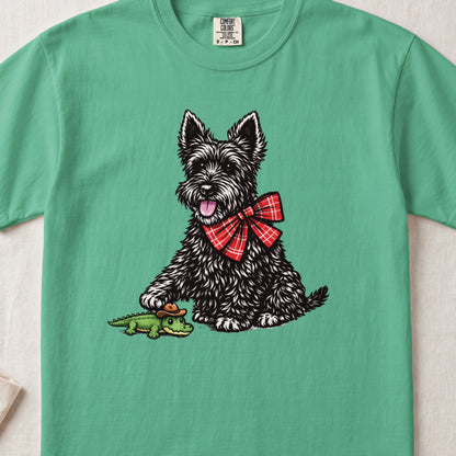 A green t-shirt printed with a black Scottish Terrier wearing a red plaid bow tie and touching a toy alligator.