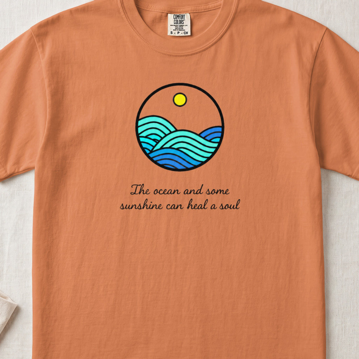 An orange t-shirt featuring a circular ocean graphic with a sun and the text "The ocean and some sunshine can heal a soul."