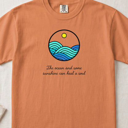 An orange t-shirt featuring a circular ocean graphic with a sun and the text "The ocean and some sunshine can heal a soul."