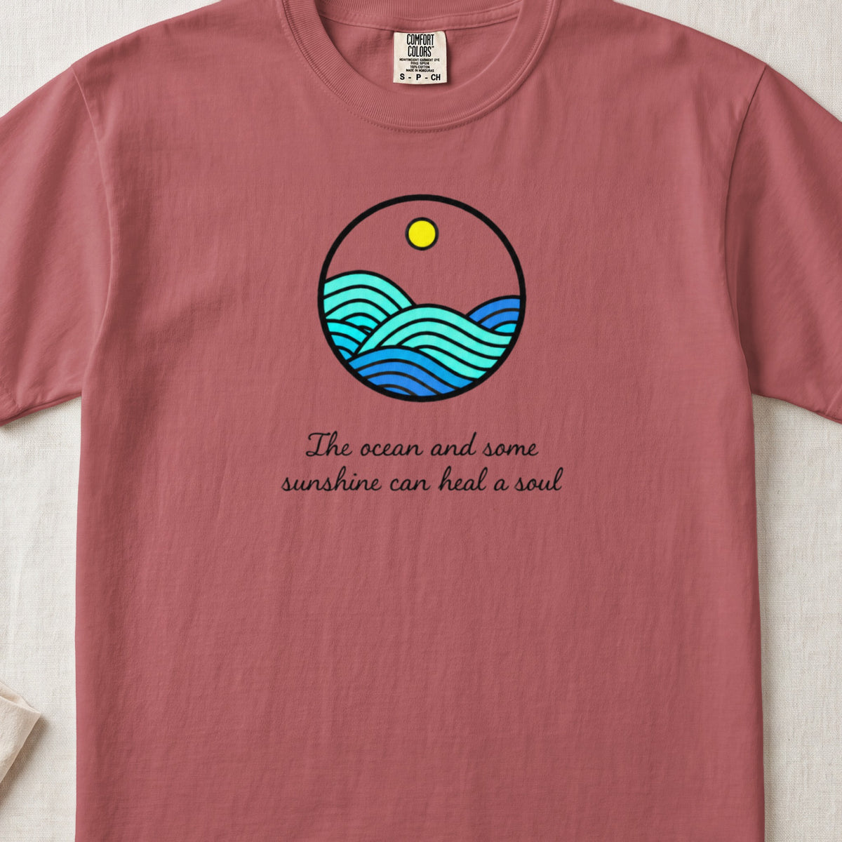 A dusty rose t-shirt featuring a circular graphic of ocean waves, a sun, and cursive text.
