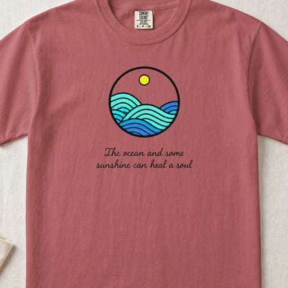 A dusty rose t-shirt featuring a circular graphic of ocean waves, a sun, and cursive text.