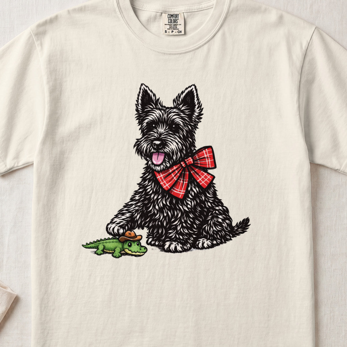 A cream-colored t-shirt with an illustration of a black terrier wearing a red plaid bow, touching a toy alligator.