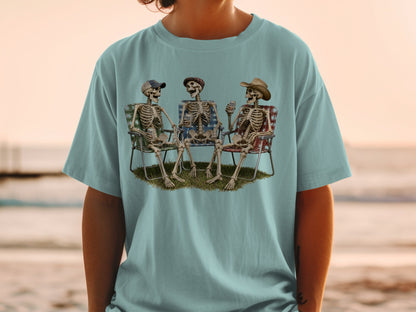 Three skeletons in lawn chairs with drinks and hats on grass.