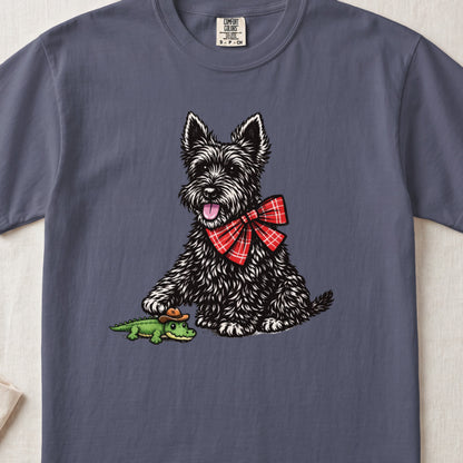 A blue crewneck t-shirt featuring an illustration of a black Scottie dog wearing a red plaid bow, touching a small toy alligator wearing a cowboy hat.