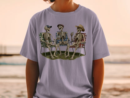 Three skeletons sit in lawn chairs on grass, holding drinks.
