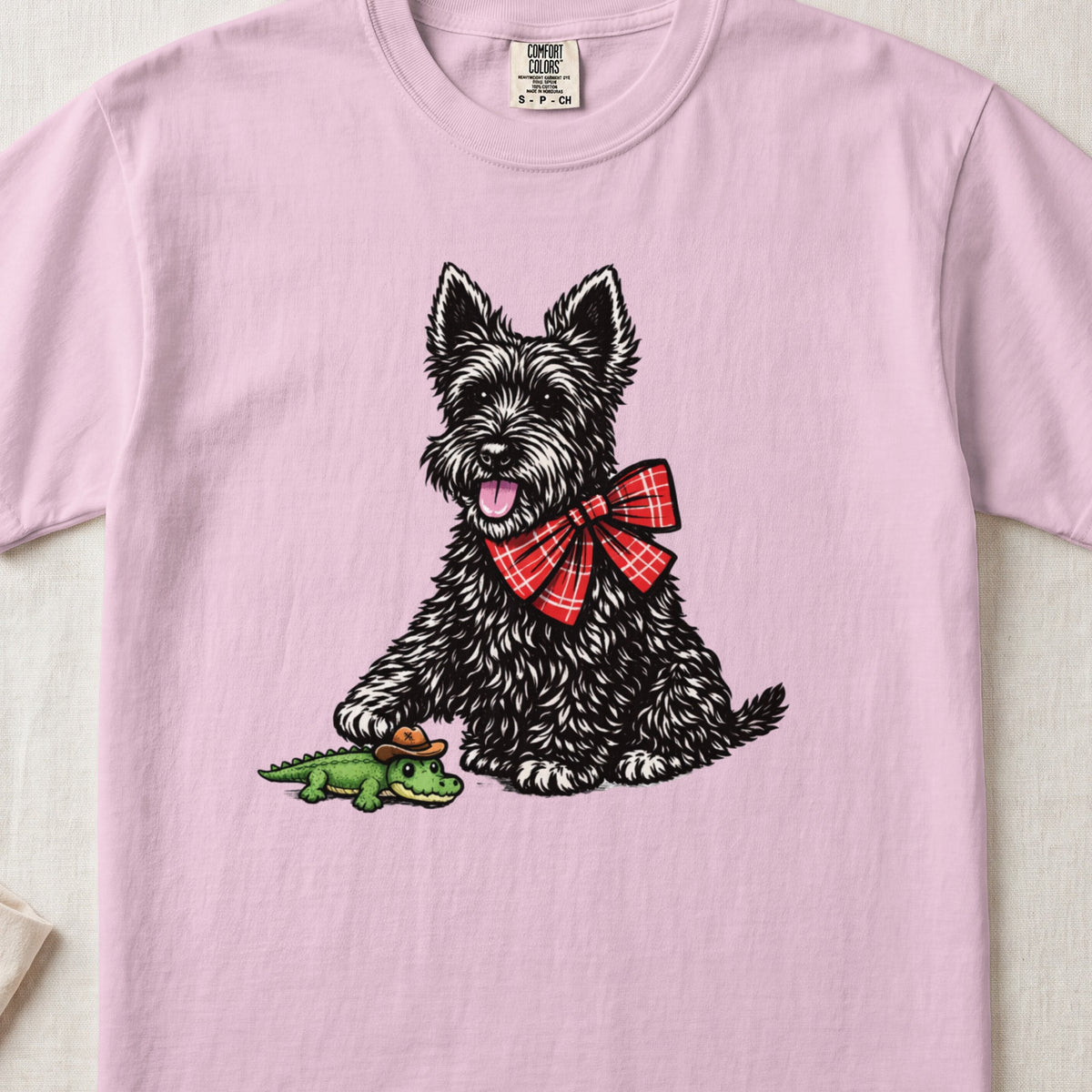A light pink t-shirt featuring an illustration of a black terrier wearing a red plaid bow, touching a toy alligator.