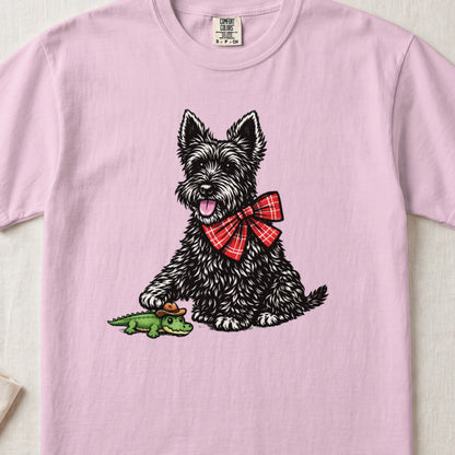 A light pink t-shirt featuring an illustration of a black terrier wearing a red plaid bow, touching a toy alligator.