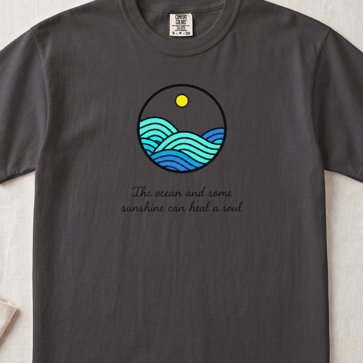 A dark gray crewneck t-shirt featuring a circular ocean graphic and the text The ocean and some sunshine can heal a soul.