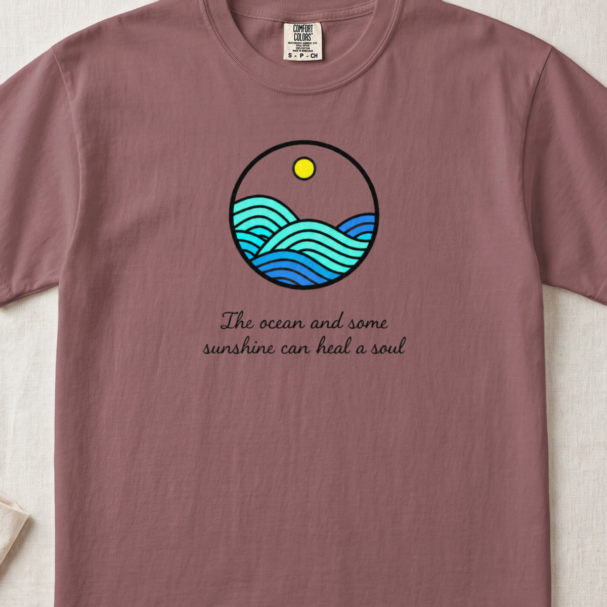 Dusty pink t-shirt featuring a circular ocean wave illustration and the text The ocean and some sunshine can heal a soul.