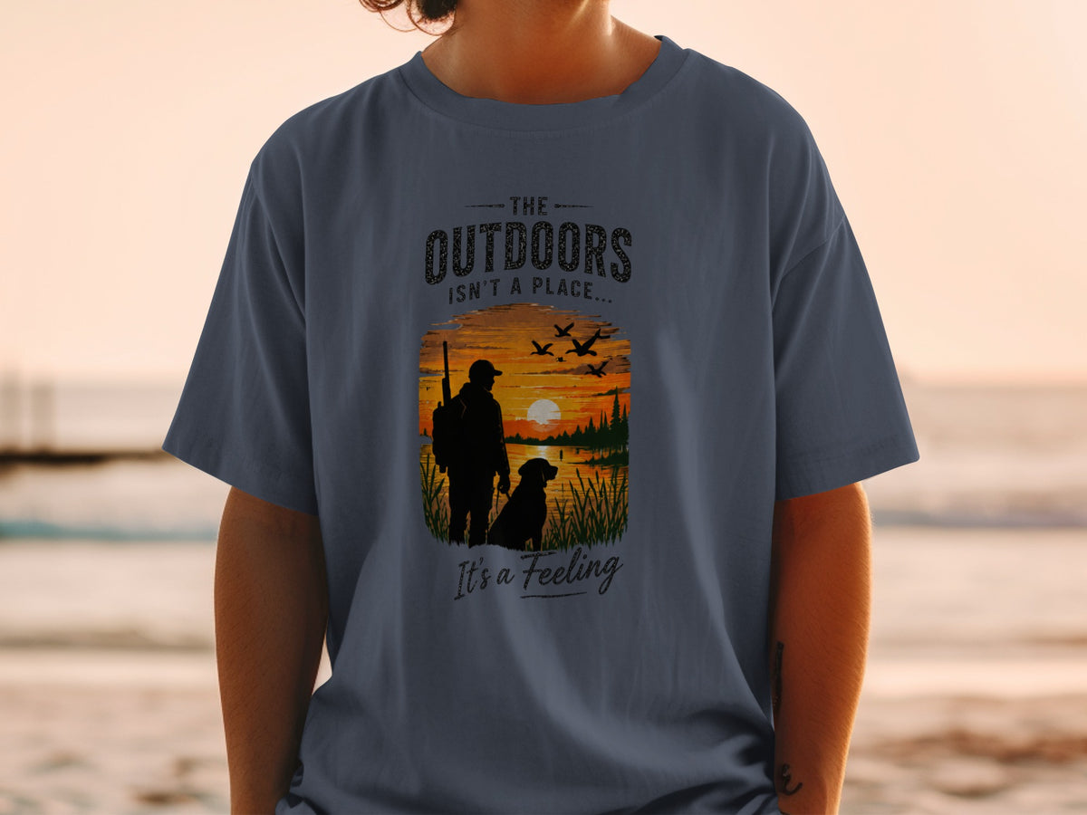 T-shirt graphic with silhouette of hunter, dog, sunset, birds, and text "The Outdoors Isn't a Place... It's a Feeling".