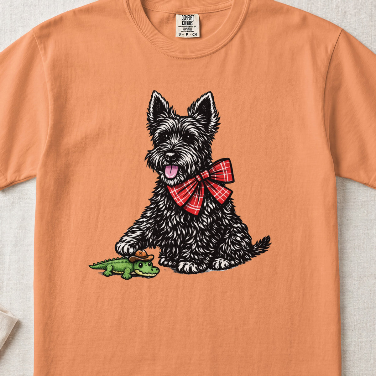 Peach-colored crewneck t-shirt featuring a graphic of a black terrier wearing a red plaid bow tie next to a toy alligator wearing a tiny cowboy hat.