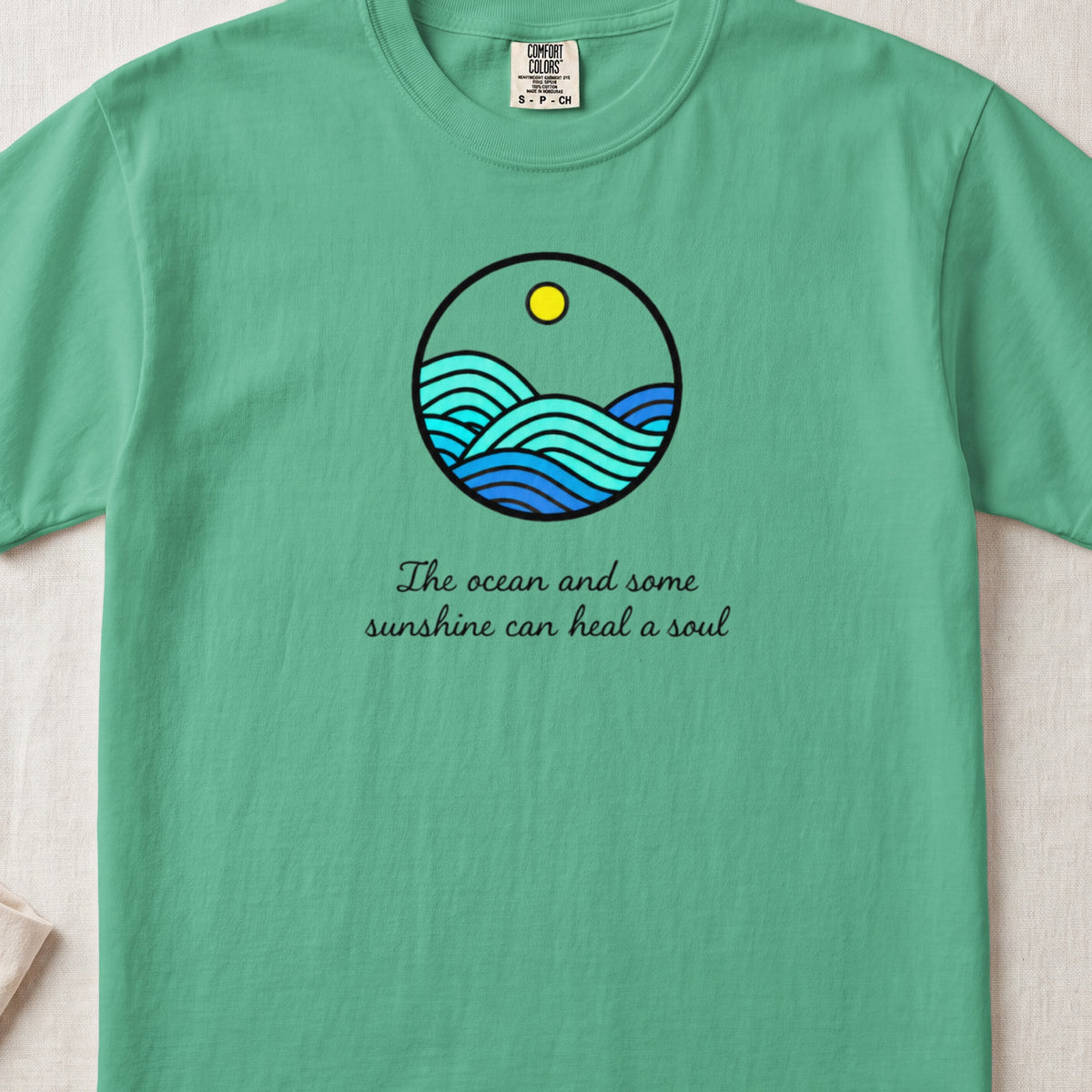 A green t-shirt featuring a circular graphic of ocean waves, a sun, and cursive text reading, "The ocean and some sunshine can heal a soul."