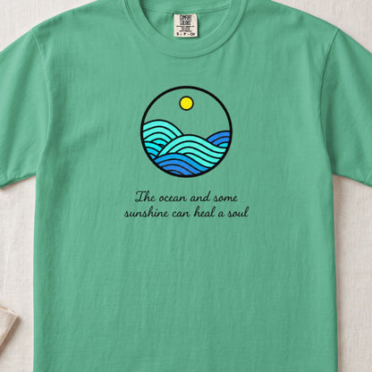 A green t-shirt featuring a circular graphic of ocean waves, a sun, and cursive text reading, "The ocean and some sunshine can heal a soul."