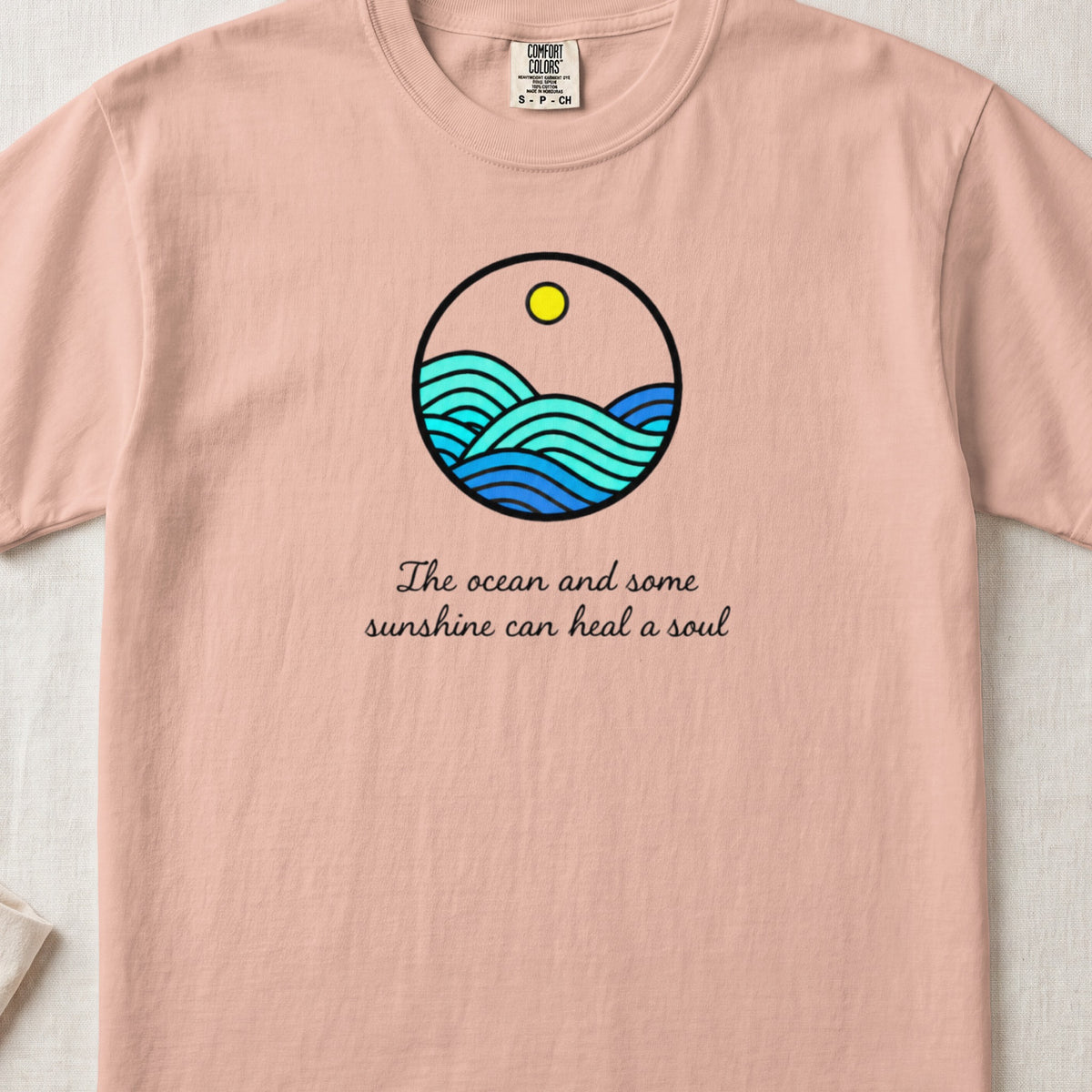 Pink t-shirt with a circular graphic of blue ocean waves, a sun, and the text The ocean and some sunshine can heal a soul.