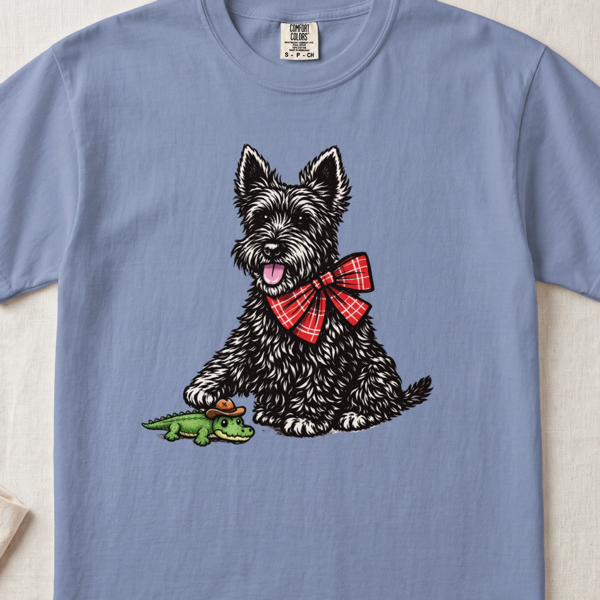 A blue crewneck t-shirt featuring an illustration of a black Scottish Terrier wearing a red plaid bow tie while holding a small toy alligator.