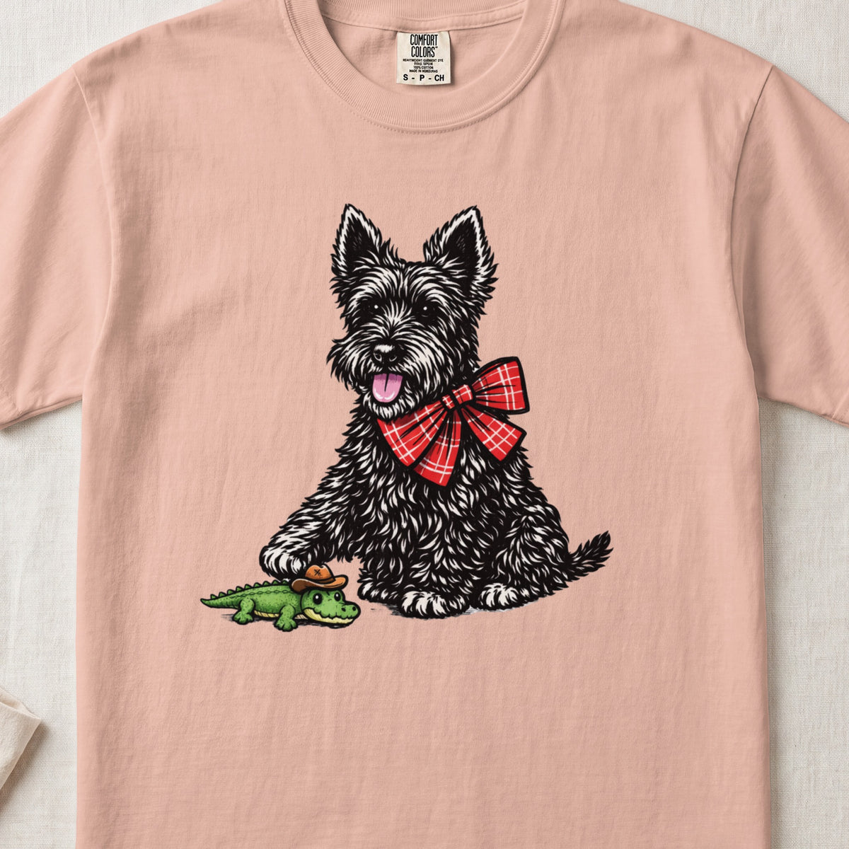 Pink t-shirt featuring an illustration of a black terrier with a red bow tie, touching a toy alligator.
