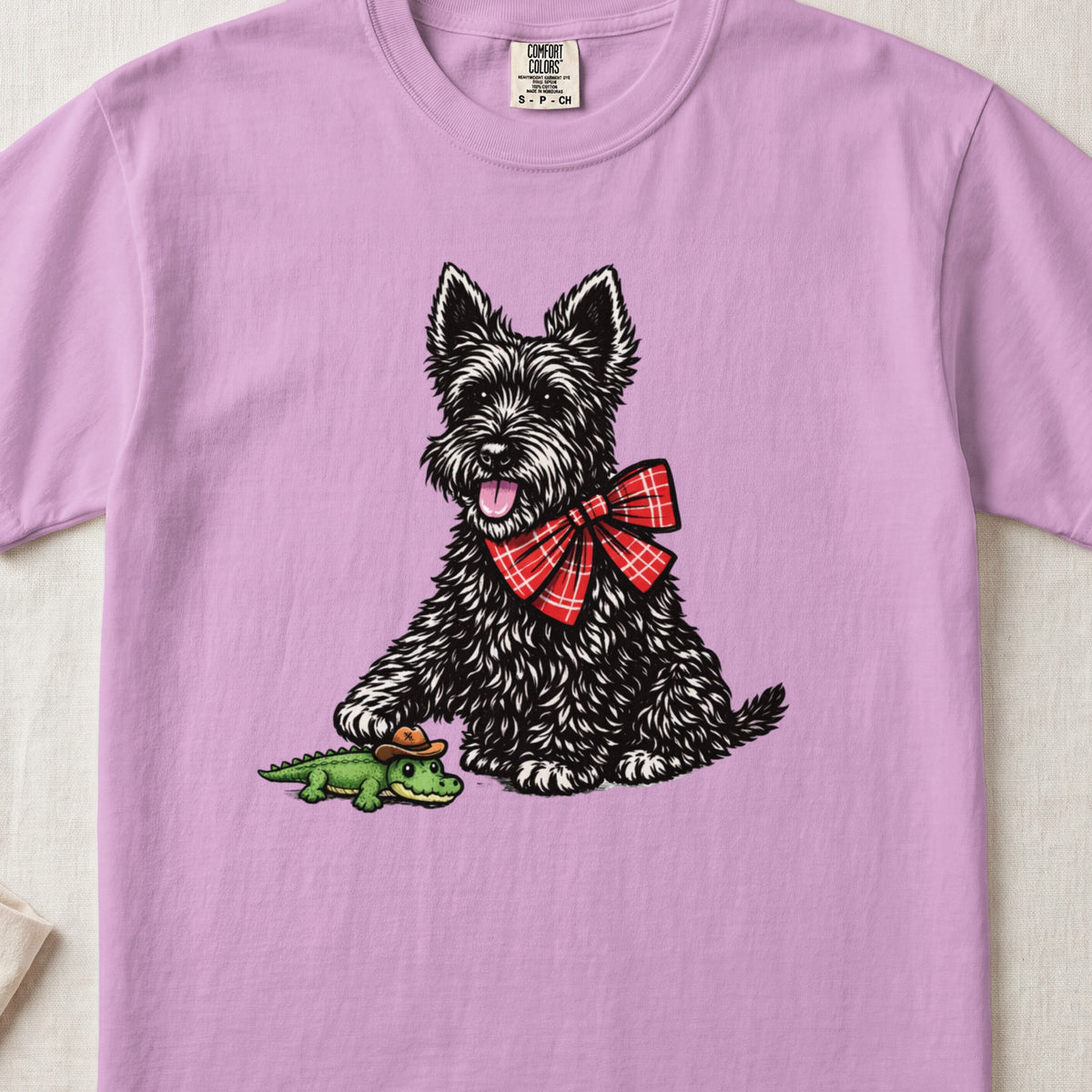 A pastel purple t-shirt features a graphic of a black terrier wearing a red plaid bow.