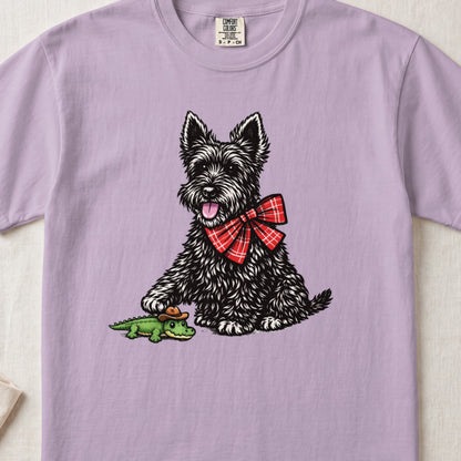A light purple t-shirt featuring a black terrier wearing a red plaid bow and touching a small toy alligator.
