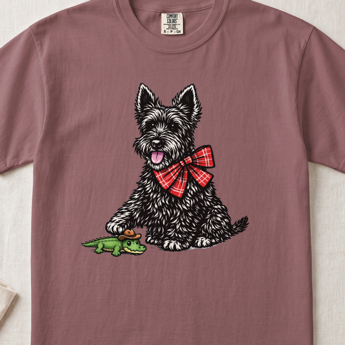 Mauve t-shirt featuring an illustration of a black Scottish Terrier wearing a red plaid bow, stepping on a toy alligator.