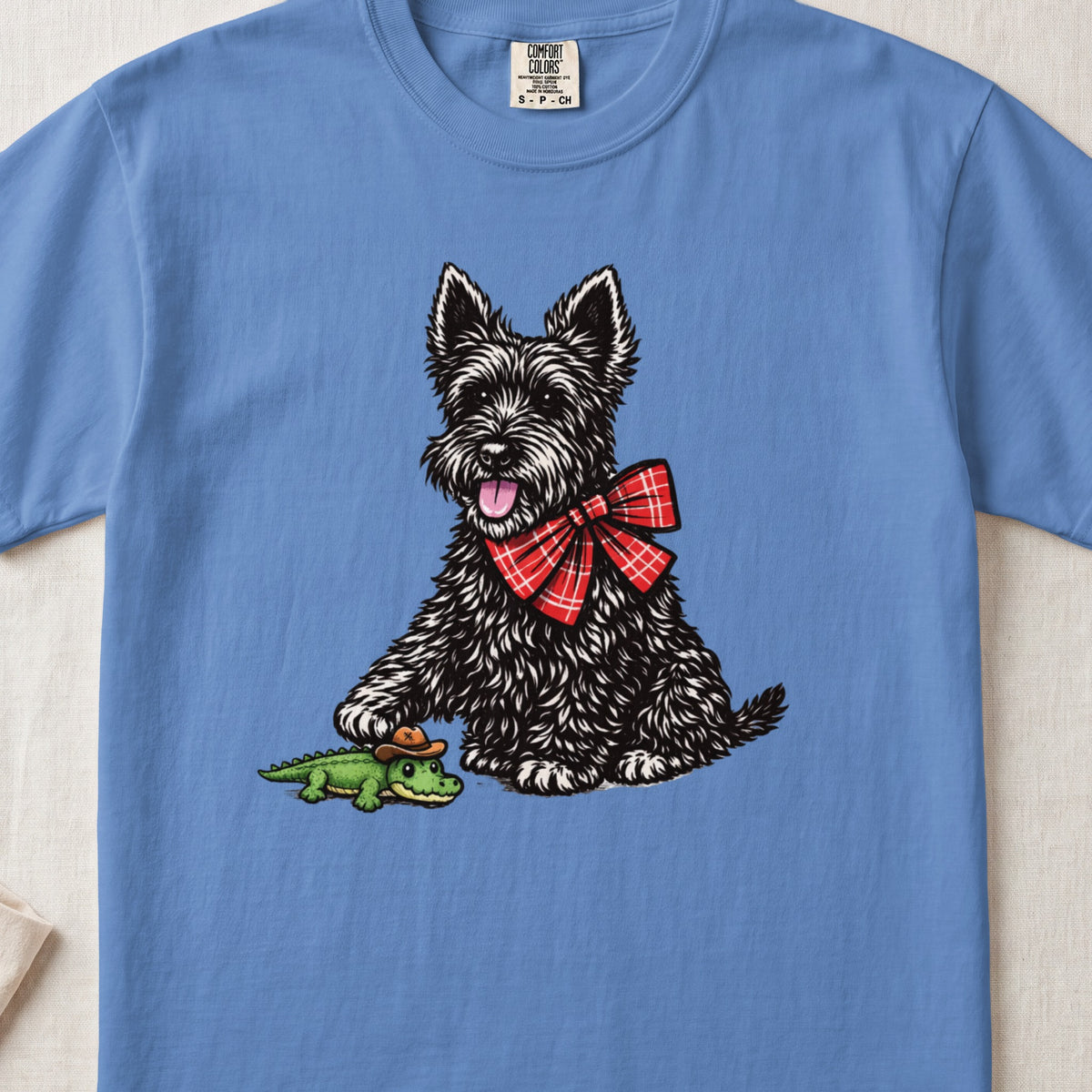 A blue t-shirt featuring a black Scottish Terrier wearing a red plaid bow tie next to a toy alligator wearing a cowboy hat.