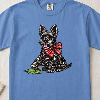 A blue t-shirt featuring a black Scottish Terrier wearing a red plaid bow tie next to a toy alligator wearing a cowboy hat.