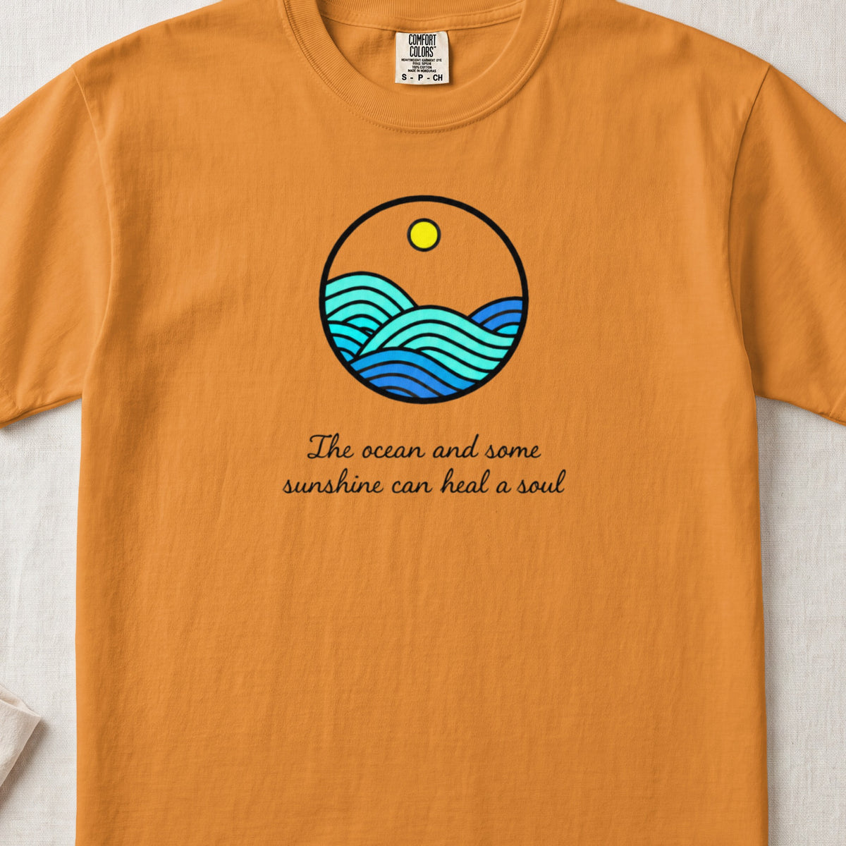 An orange t-shirt featuring a circular ocean and sunshine graphic with the text The ocean and some sunshine can heal a soul.