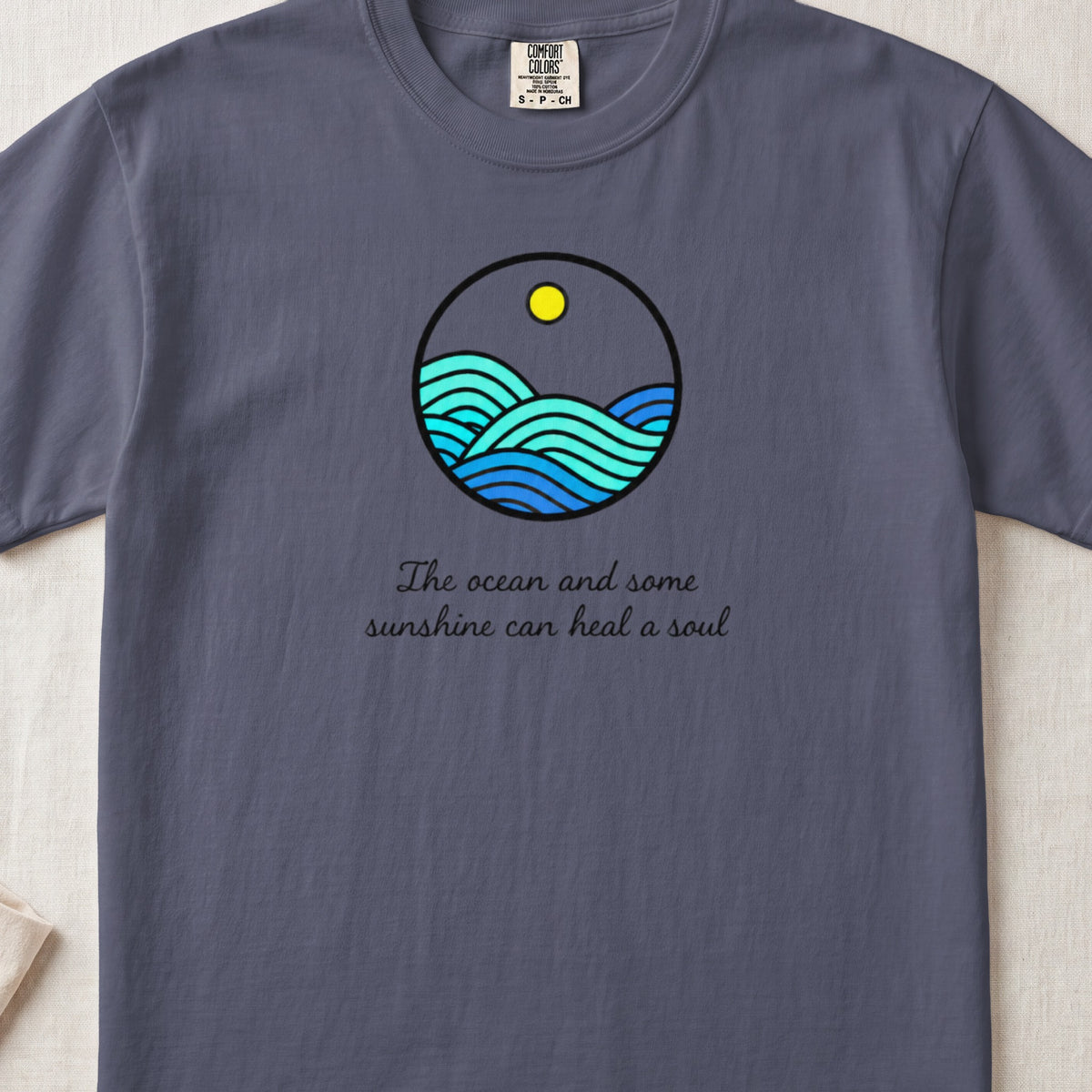 A slate grey t-shirt featuring a circular graphic of blue ocean waves, a yellow sun, and the text The ocean and some sunshine can heal a soul.