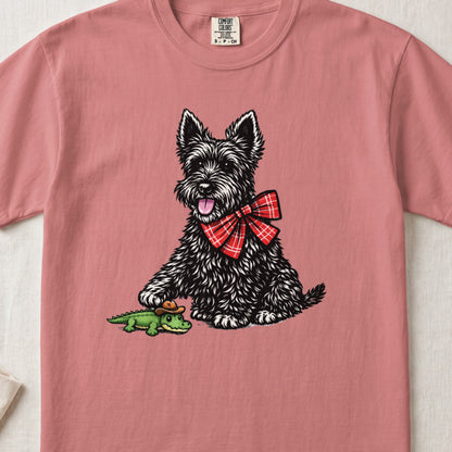 A pink t-shirt featuring a black Scottish Terrier wearing a red plaid bow tie and touching a small toy alligator.