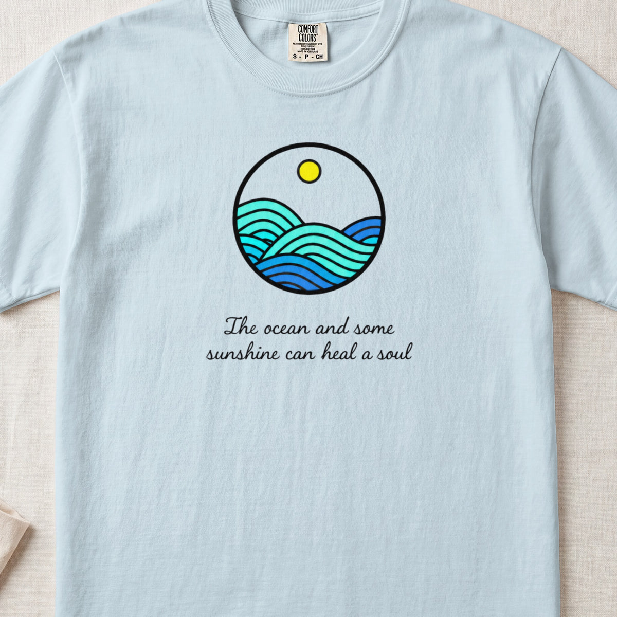 A light blue t-shirt with a round graphic of ocean waves, a sun, and cursive text.