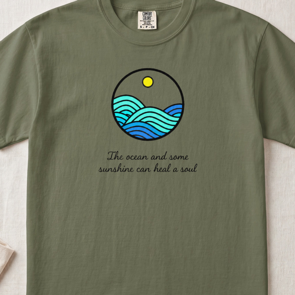 An olive green t-shirt with a round graphic of ocean waves and text reading The ocean and some sunshine can heal a soul.