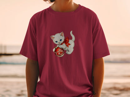 White kitten with blue eyes and red bow holding Christmas ornament on burgundy shirt.