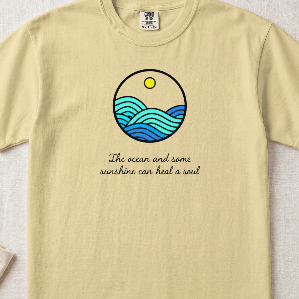 A light yellow t-shirt features a circular graphic of blue ocean waves under a yellow sun, with cursive text reading "The ocean and some sunshine can heal a soul".