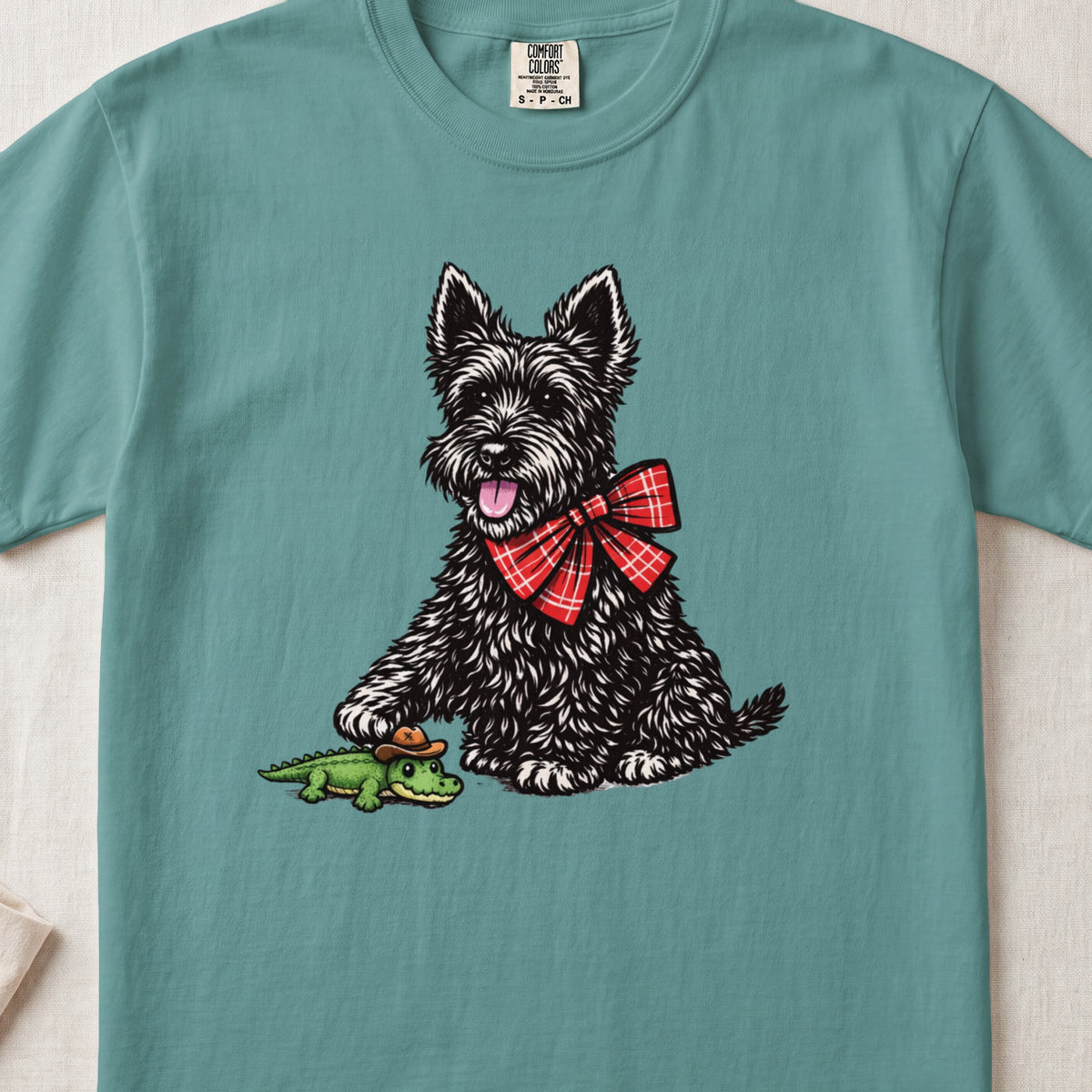 Teal t-shirt featuring an illustration of a black terrier wearing a red plaid bow tie and touching a small toy alligator.