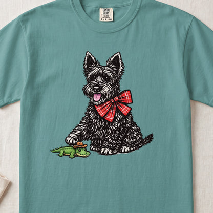 Teal t-shirt featuring an illustration of a black terrier wearing a red plaid bow tie and touching a small toy alligator.