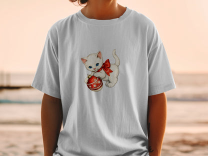 White kitten with blue eyes and red bow holding a Christmas ornament on a t-shirt.