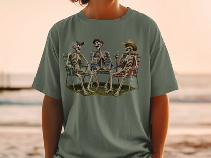 Three skeletons sit in camping chairs on grass, holding drinks, on a t-shirt.