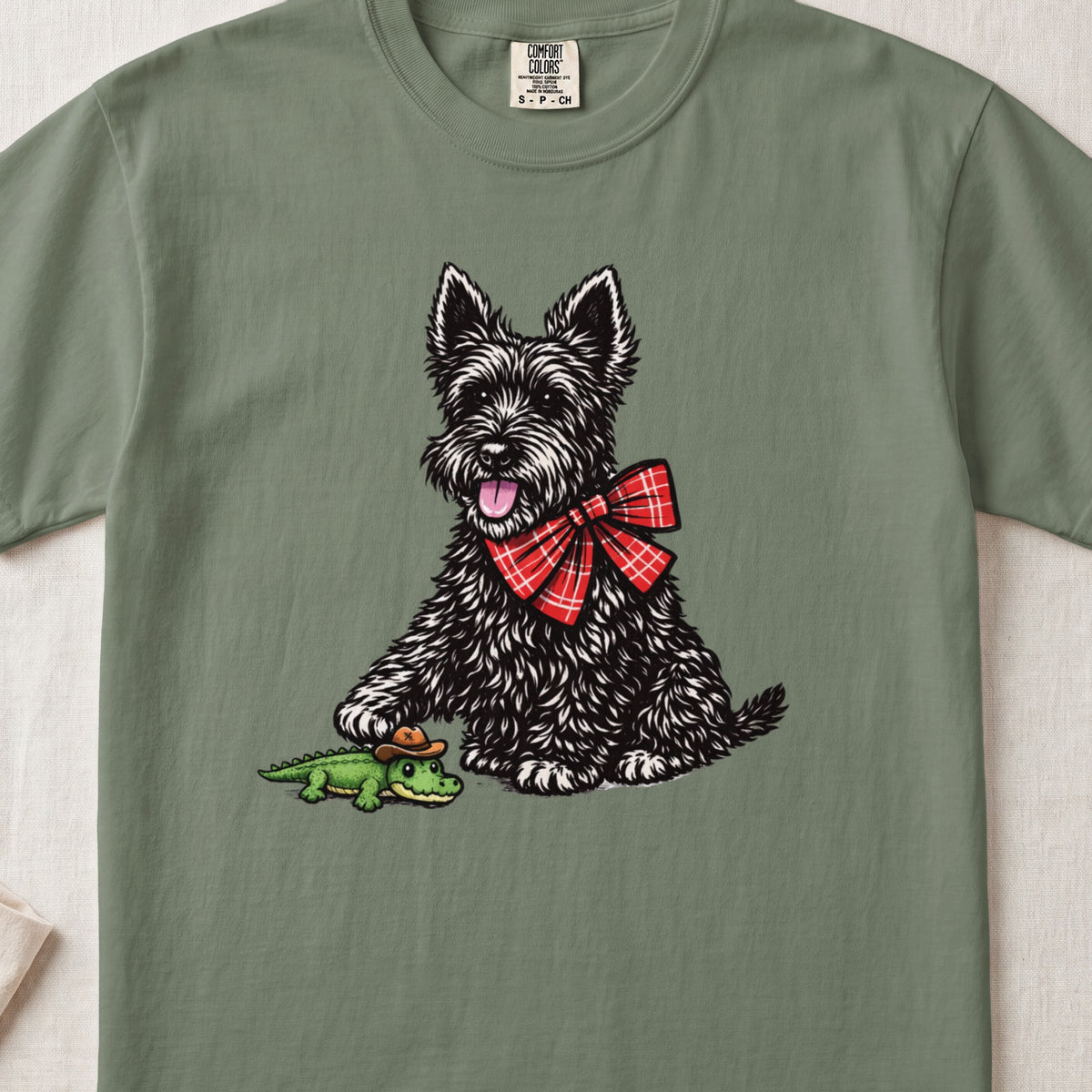 A sage green t-shirt featuring a graphic of a black terrier wearing a red plaid bow.