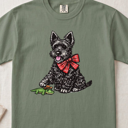 A sage green t-shirt featuring a graphic of a black terrier wearing a red plaid bow.
