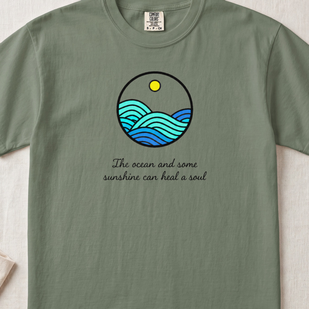Sage green t-shirt with a round graphic of ocean waves and text reading The ocean and some sunshine can heal a soul.
