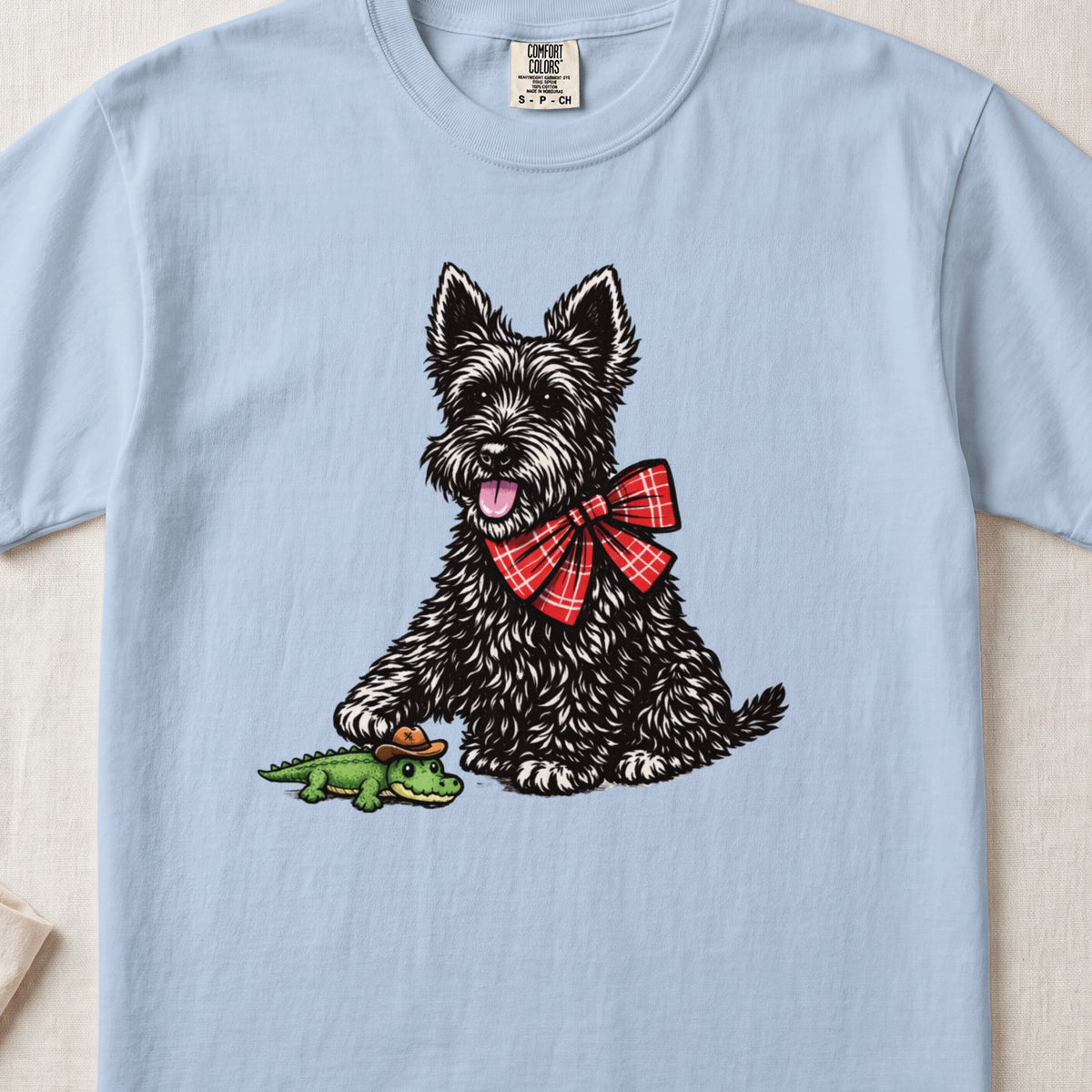 A light blue t-shirt featuring an illustration of a black terrier wearing a red plaid bow, touching a tiny green alligator wearing a cowboy hat.