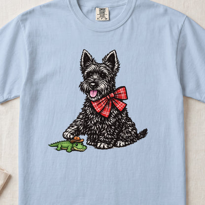 A light blue t-shirt featuring an illustration of a black terrier wearing a red plaid bow, touching a tiny green alligator wearing a cowboy hat.