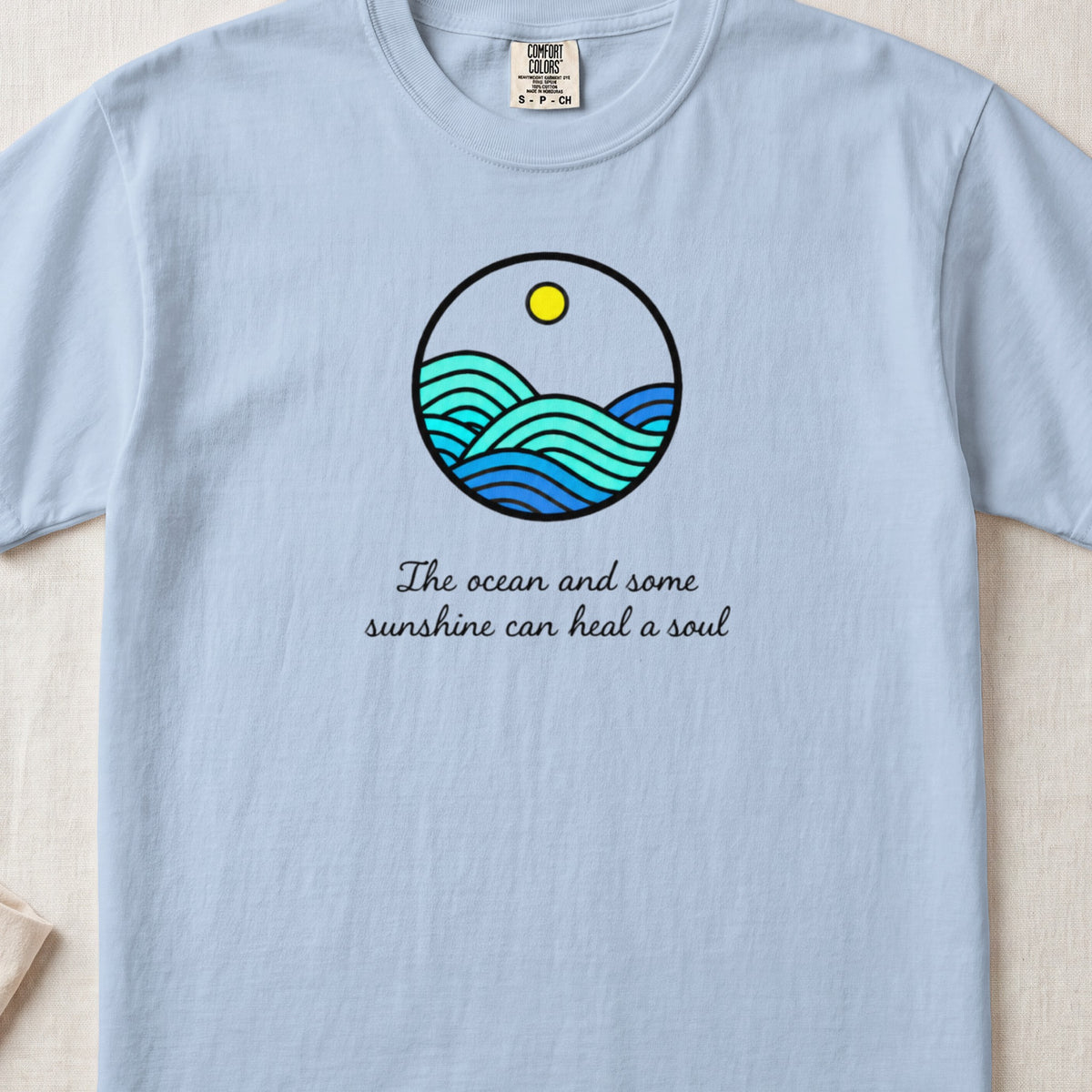 A light blue t-shirt with a round graphic of ocean waves and text reading The ocean and some sunshine can heal a soul.
